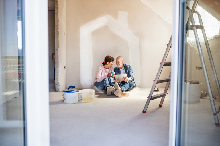 Paar sitzt auf dem Boden eines unfertigen Zimmers mit einer Hausumrisszeichnung an der Wand, umgeben von Farbeimern und einer Leiter