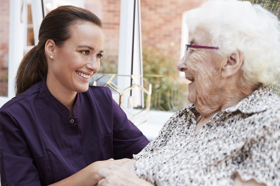 Eine Altenbetreuung kümmert sich um eine Seniorin, die in ihrem Sessel sitzt.