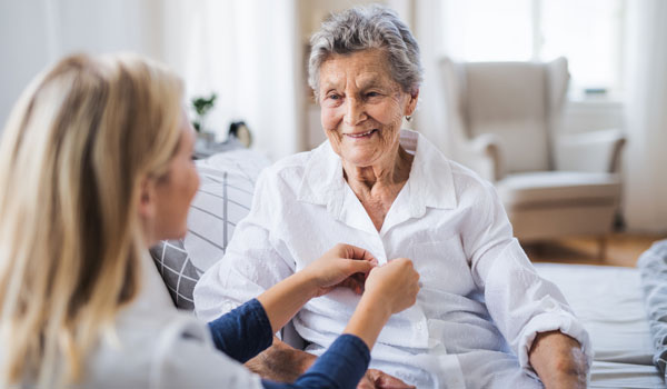 Eine Altenbetreuerin hilft einer älteren Seniorin ihre Knöpfe an der Bluse zu zumachen.