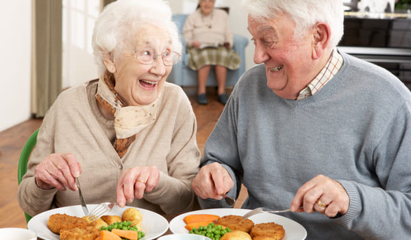 Ein Seniorenpaar sitzt am Mittagstisch und ißt ein Schnitzel mit Kartoffeln, Erbsen und Möhren.