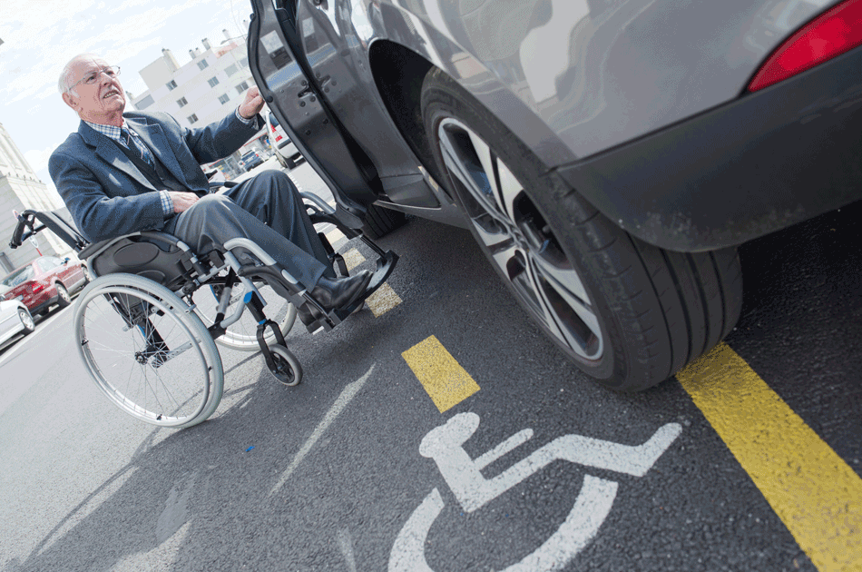 Ein Rollstuhlfahrer vor seinem Auto, welches auf einem Behindertenparkplatz steht.