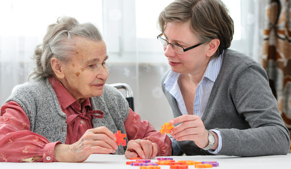 Eine Seniorin spielt mit einem Puzzle und wird dabei von einer jüngeren Frau unterstützt.