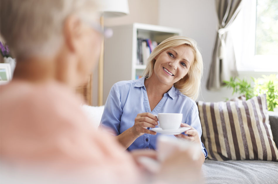Ein Besuchsdienst besucht eine ältere Dame. Beide siitzen im Wohnzimmer und trinken Kaffee.