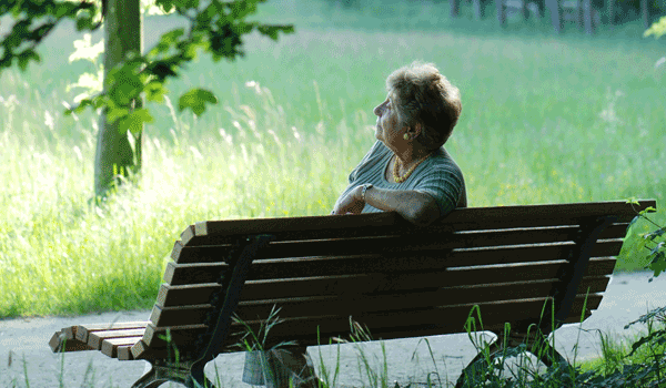Eine ältere Frau sitzt auf einer Parkbank und lauscht den Geräuschen im Park.