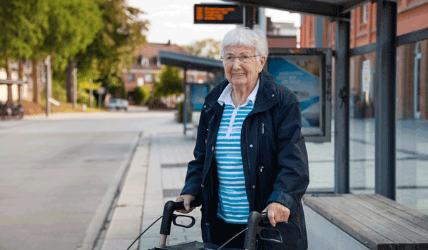Eine Seniorin mit Rollator wartet an der Haltestelle auf den Bus.