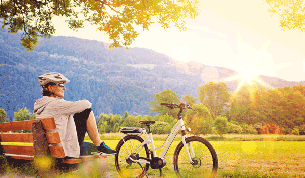 Ein Radwanderin macht Pause. Sie sitzt auf der Bank und Ihr Fahrrad hat sie vor sich abgestellt. Dabei blickt sie auf die tiefscheinende Sonne.