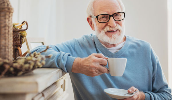 Senior steht entspannt im Wohnzimmer mit einer Tasse Kaffee in der Hand.