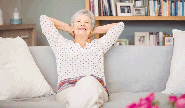 Eine Senioren sitzt auf der Cocuh, hat ihre Arme hinter ihrem Kopf verschränkt, und blickt zufrieden drein.