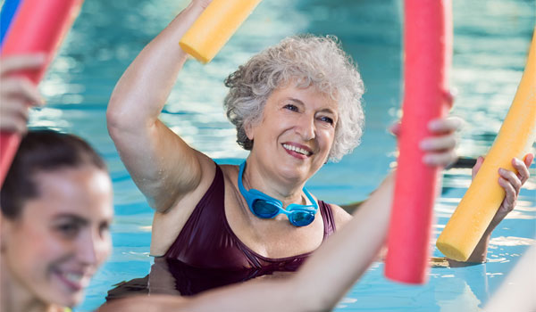 Seniorin bei der Wassergymnastik. Sie hält eine Schwimmnudel über den Kopf und lächelt dabei.