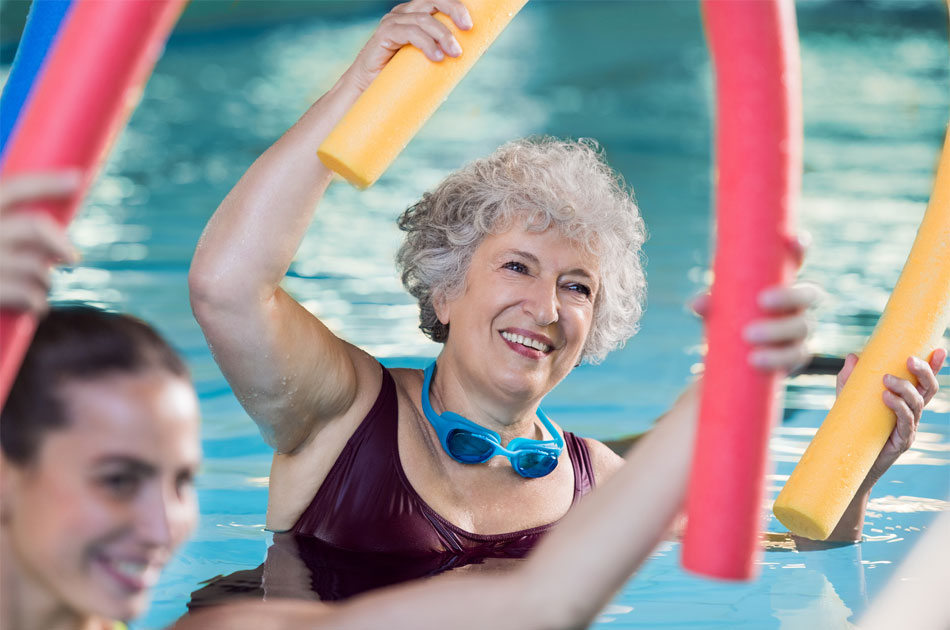 Seniorin bei der Wassergymnastik. Sie hält eine Schwimmnudel über den Kopf und lächelt dabei.