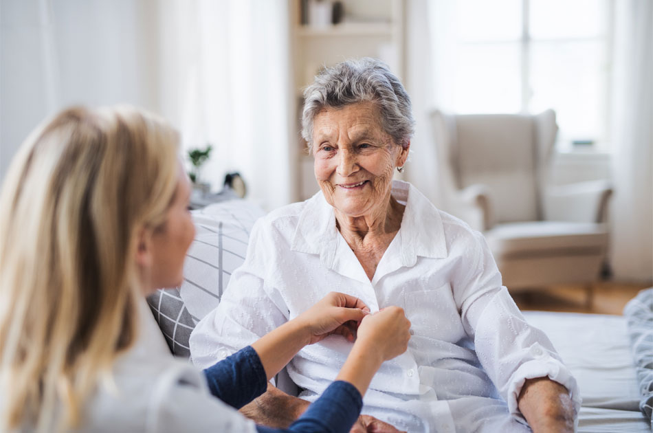 Eine Ersatzpflegekraft führt Verhinderungspflege bei einer Seniorin durch, die auf ihrem Bett sitzt, während die Pflegerin ihre Jacke zuknöpft.