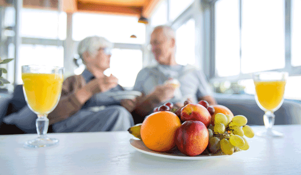 Ein Seniorenpaar auf der Couch. Vor ihnen auf dem Tisch steht ein Teller Obst und zwei Gläser Saft.
