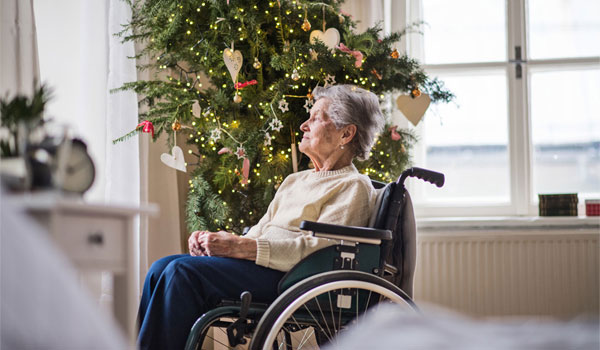 Eine Frau sitzt einsam im Rollstuhl und schaut gedankenverloren aus dem Fenster., neben ihr steht ein geschmückter Weihnachtsbaum.