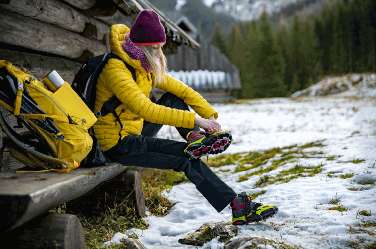 Eine Frau sitzt auf einer Bank vor einer Holzhütte. Es ist Winter und sie zieht sich ihre Wanderschuhe an.