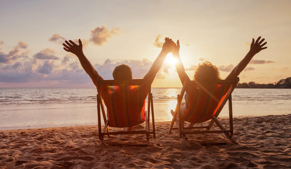 Ein Rentnerpaar sitzt auf ihren Liegestühlen und schauen sich den Sonnenuntergang am Meer an. Dabei heben sie ihre Arme.