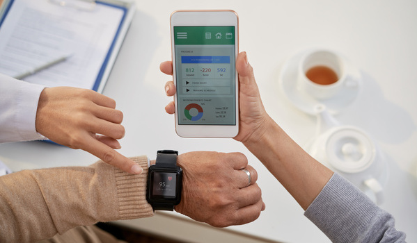 Sein Senior schaut auf seine Smart Watch und überprüft seine Daten auf dem Handy. Dabei wird er von einer Ärztin unterstützt.