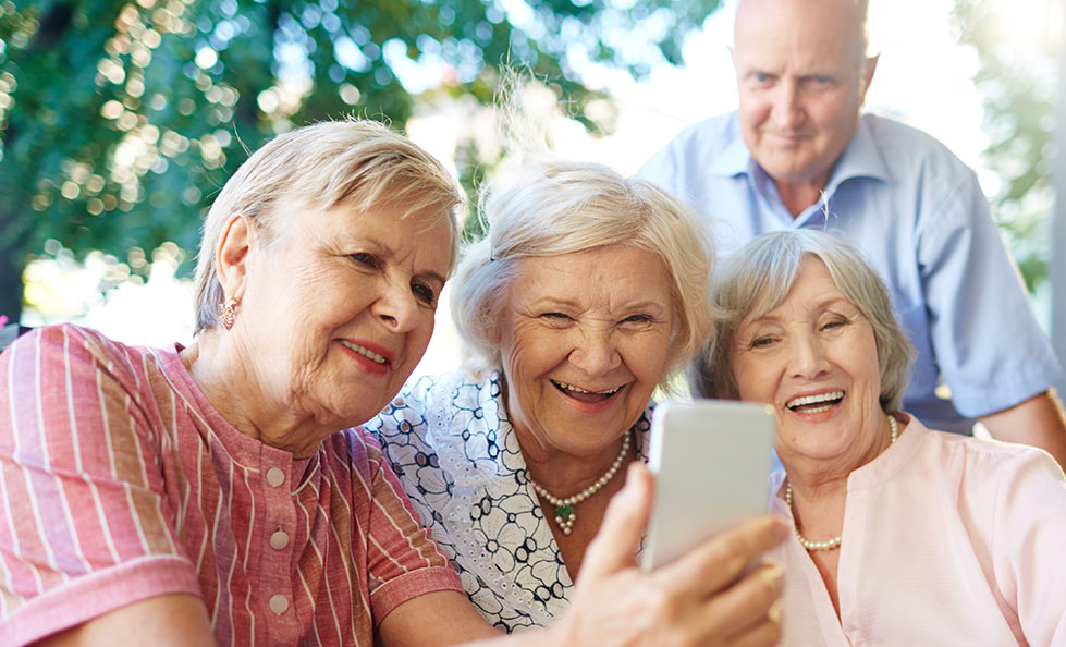 Eine Seniorengruppe aus drei Frauen und einem Mann schauen auf ein Handy.