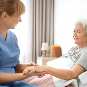 Pflegekraft im blauen Kittel hält die Hände einer älteren Patientin im Krankenhausbett