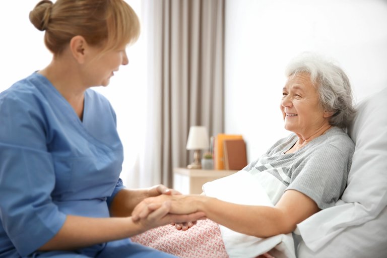 Pflegekraft im blauen Kittel hält die Hände einer älteren Patientin im Krankenhausbett