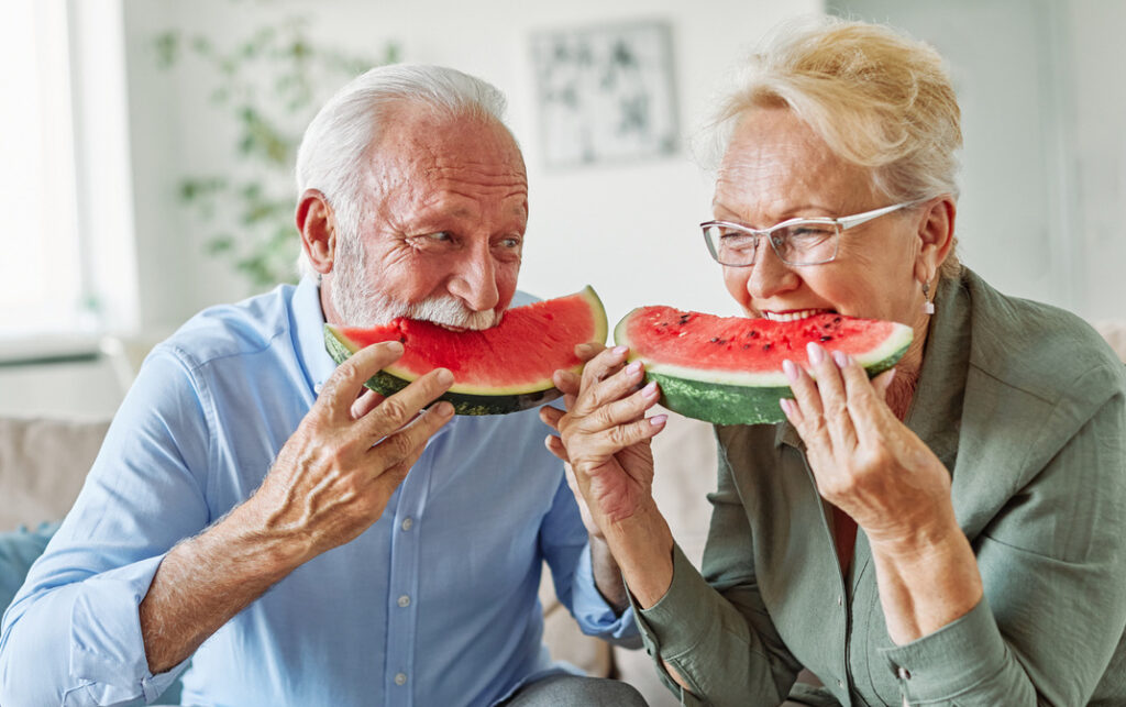 Porträt eines glücklichen älteren Paares, das sich umarmt, Wassermelone isst und zu Hause Spaß hat.