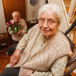 Eine Seniorin fährt mit dem Treppenlift die Treppe hoch. Im Hintergrund ist ihr Mann mit Rosen in der Hand zu sehen.
