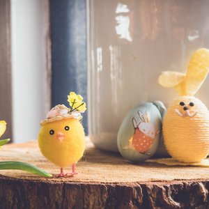Gelbes Küken mit Blumenhut, bemaltes Osterei mit Hasenmotiv und gelber Osterhase aus Garn auf Holzscheibe neben Narzisse