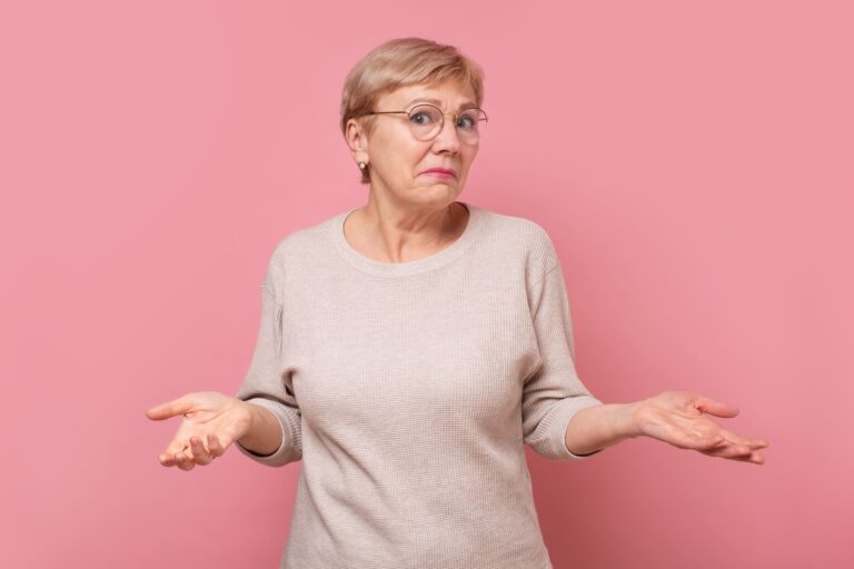 Portrait einer älteren Frau, die fragend vor einem rosa Hintergrund steht.